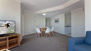 Un salon avec un téléviseur, une table à manger avec des chaises, un canapé bleu et, au fond, un coin cuisine.