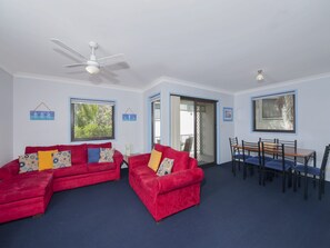 A room with two red sofas, a ceiling fan, a dining table with chairs, and a view of greenery outside. - Carindale 21 Nelson Bay Apartment with Pool (Nelson Bay)
