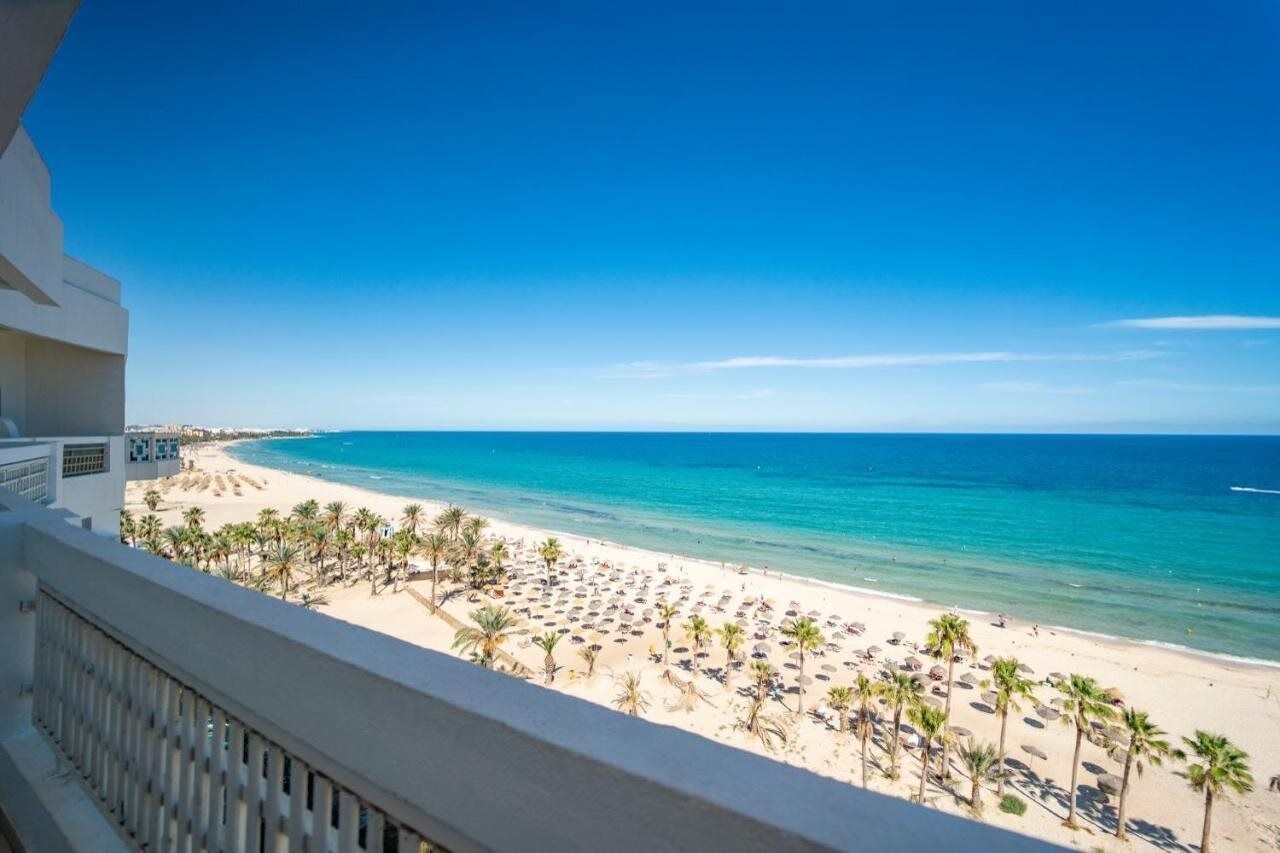 A beach view from a balcony with palm trees and umbrellas.