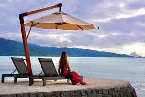 A woman seated on a lounge chair under a canopy, overlooking a body of water with mountains in the distance. - EDEN - Private Island - TAHA'A (Taha'a)
