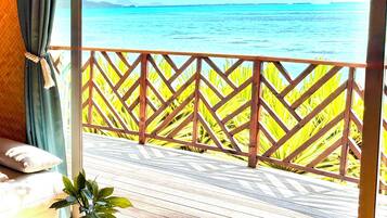 A room with a wooden deck overlooking a turquoise sea, a view of distant land, and a tropical plant on the floor.