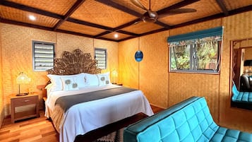 Un dormitorio con cama, cabecero tapizado azul, ventilador de techo y ventana con vista a follaje.