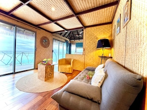 Un salón amplio con suelo de madera, un sofá, una butaca, un tapete redondo y un ventanal con vistas al mar.