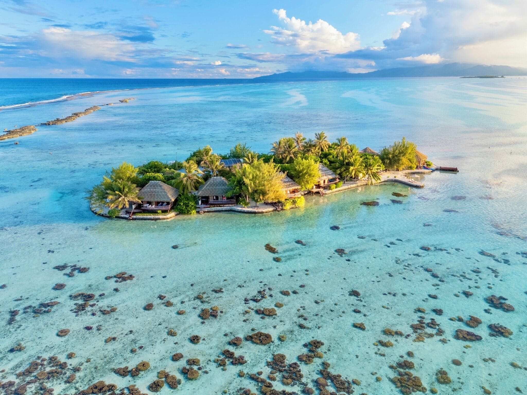 An overwater bungalow resort with thatched-roof structures, clear turquoise waters, and a small island surrounded by palm trees.