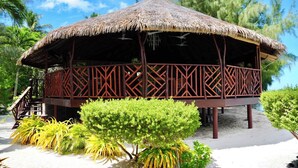 A thatched-roof structure with a wooden lattice design, situated on a sandy beach with tropical vegetation.