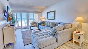 A modern living room with a grey sectional sofa, a white side table, and a lamp. There is a large window with a view of the sea. - Villa Madeira 203 Gorgeous remodel|Walk to John's Pass|Large Balcony! (Madeira Beach)