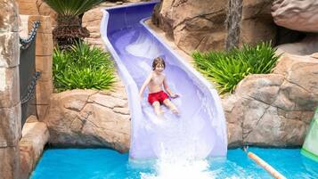 Un enfant glisse sur un toboggan aquatique et atterrit dans une piscine entourée de rochers et de plantes.