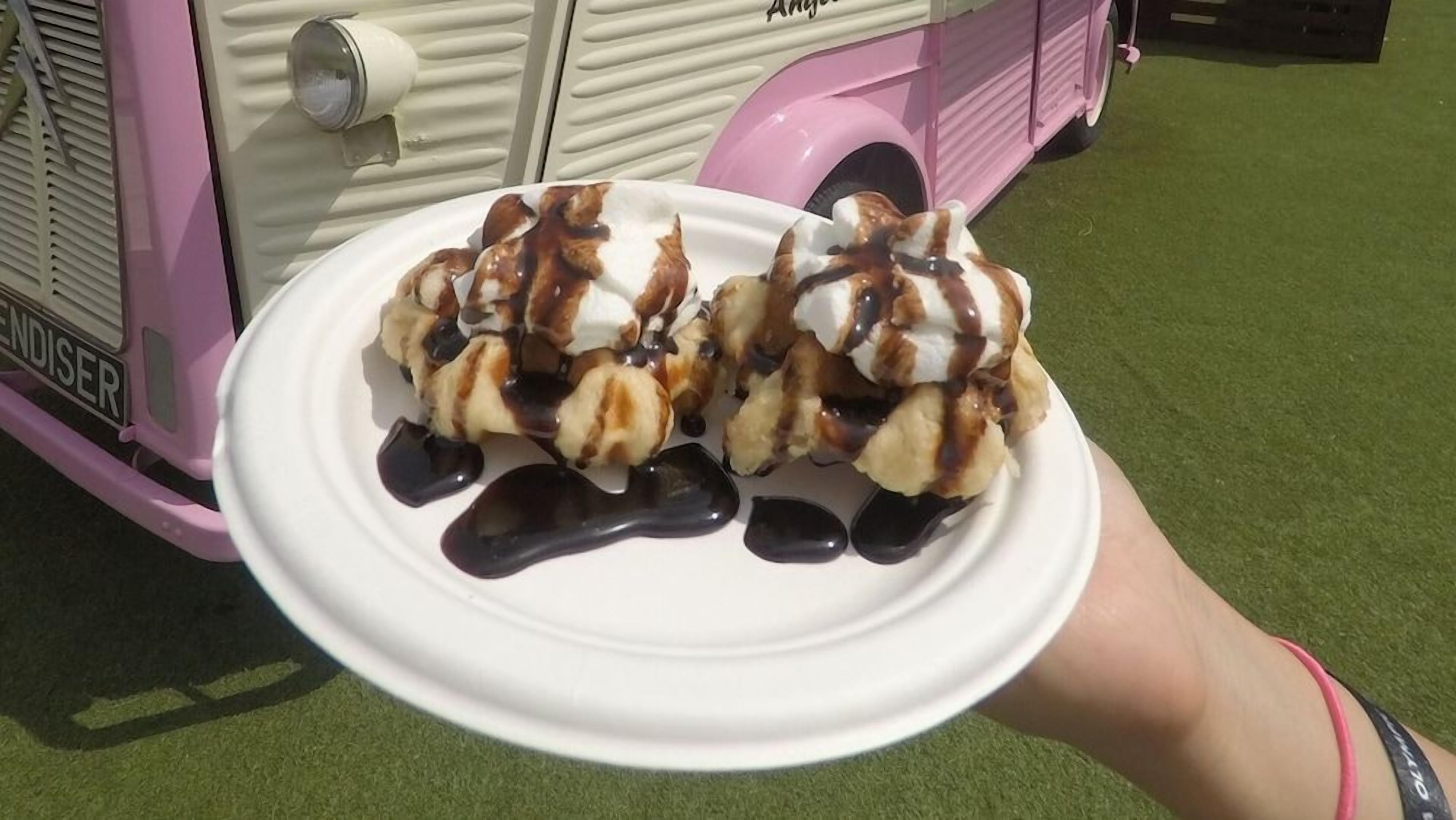 Un plato blanco con dos postres cubiertos de crema batida y salsa de chocolate, sostenido por una persona con una pulsera rosa.