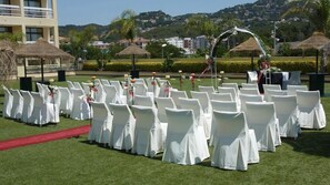 A wedding ceremony setup with rows of white chairs and a red carpet on a grassy lawn, with a backdrop of buildings and hills. - Evenia Olympic Resort - Olympic Park (Lloret de Mar)