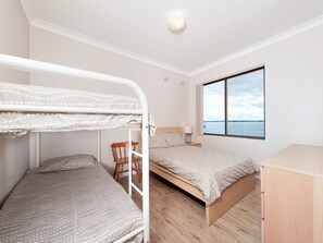 Een kamer met stapelbedden, een houten stoel, een nachtkastje met een lamp en een raam met uitzicht op zee.