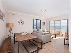 A modern living room with a wooden floor, a sofa, a dining table, and a view of the sea through large windows.