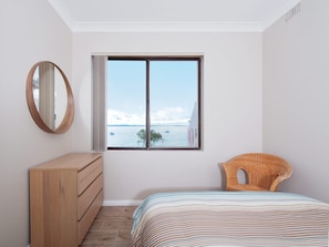 Een slaapkamer met een groot raam, een houten dressoir, een rieten stoel en een gestreept dekbed.
