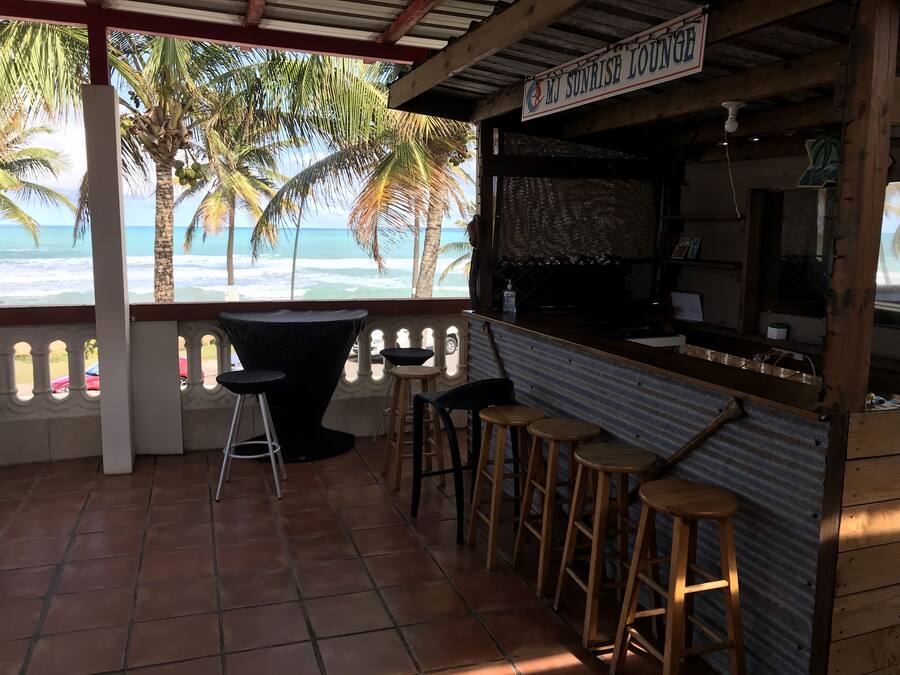 A seaside lounge with a view of the ocean, featuring a bar area with stools and a table, and a covered seating area with a view of palm trees.
