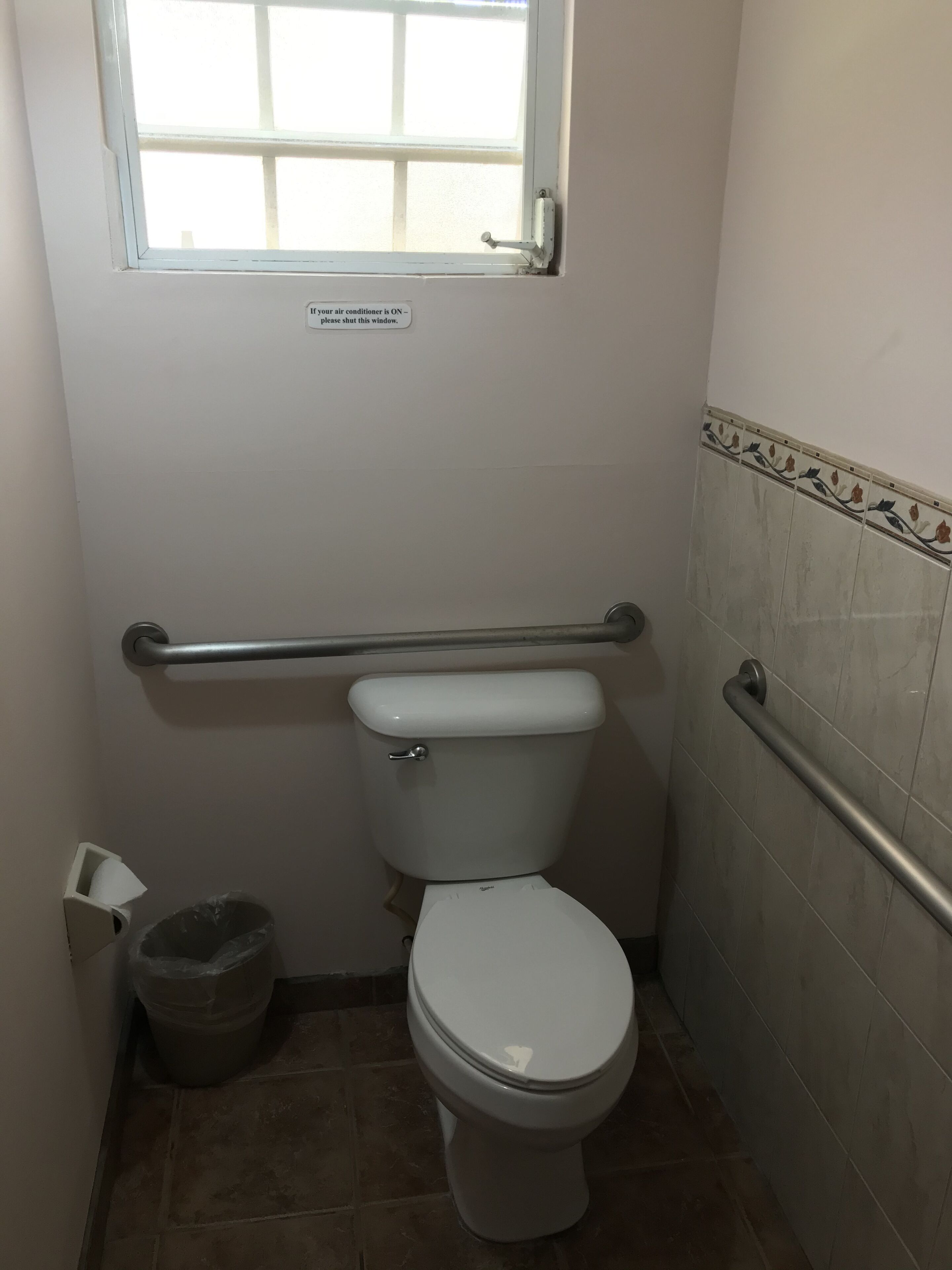 Ein kleines Badezimmer mit Toilette, Handlauf, Fenster und Mülleimer.