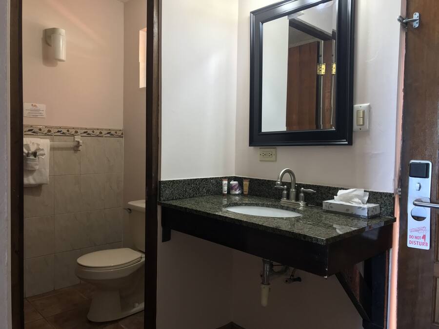 Un baño con piso de loseta de mármol, inodoro blanco, lavamanos con cubierta de granito, un espejo y dispensador de toallas.