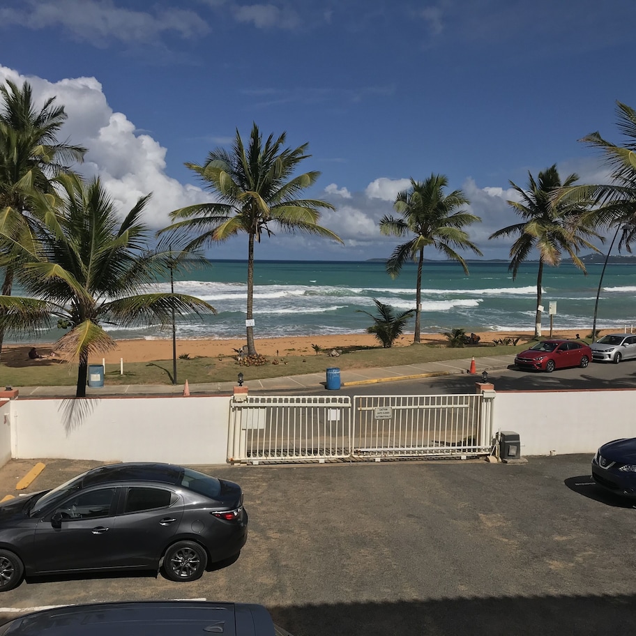 A parking area with cars, a gated entrance, and a view of the beach with palm trees and ocean waves.