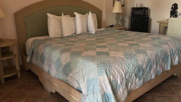 Un dormitorio con cama, mesita de noche, lámpara, microondas y una pequeña mesa con cafetera.