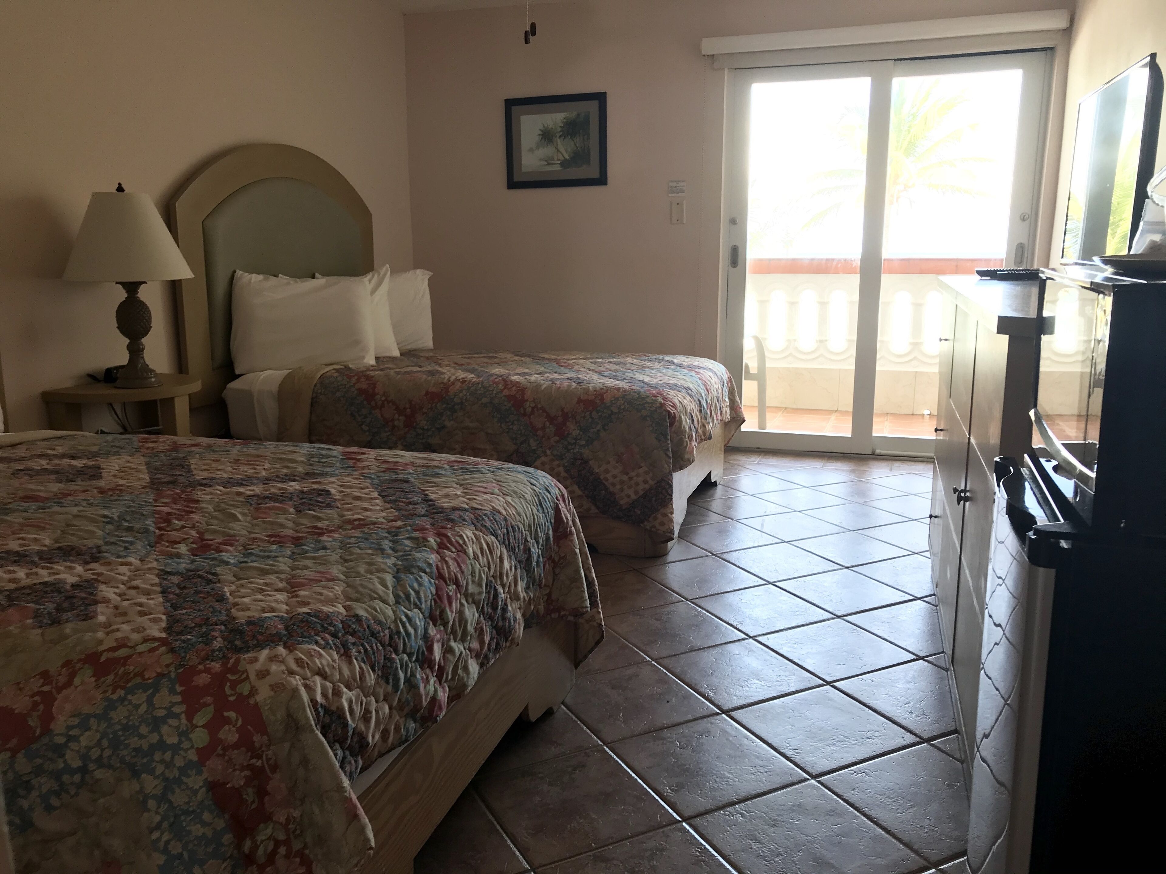 Habitación de hotel con dos camas, una mesita de noche, una lámpara, un televisor y una puerta corrediza de vidrio hacia un balcón.