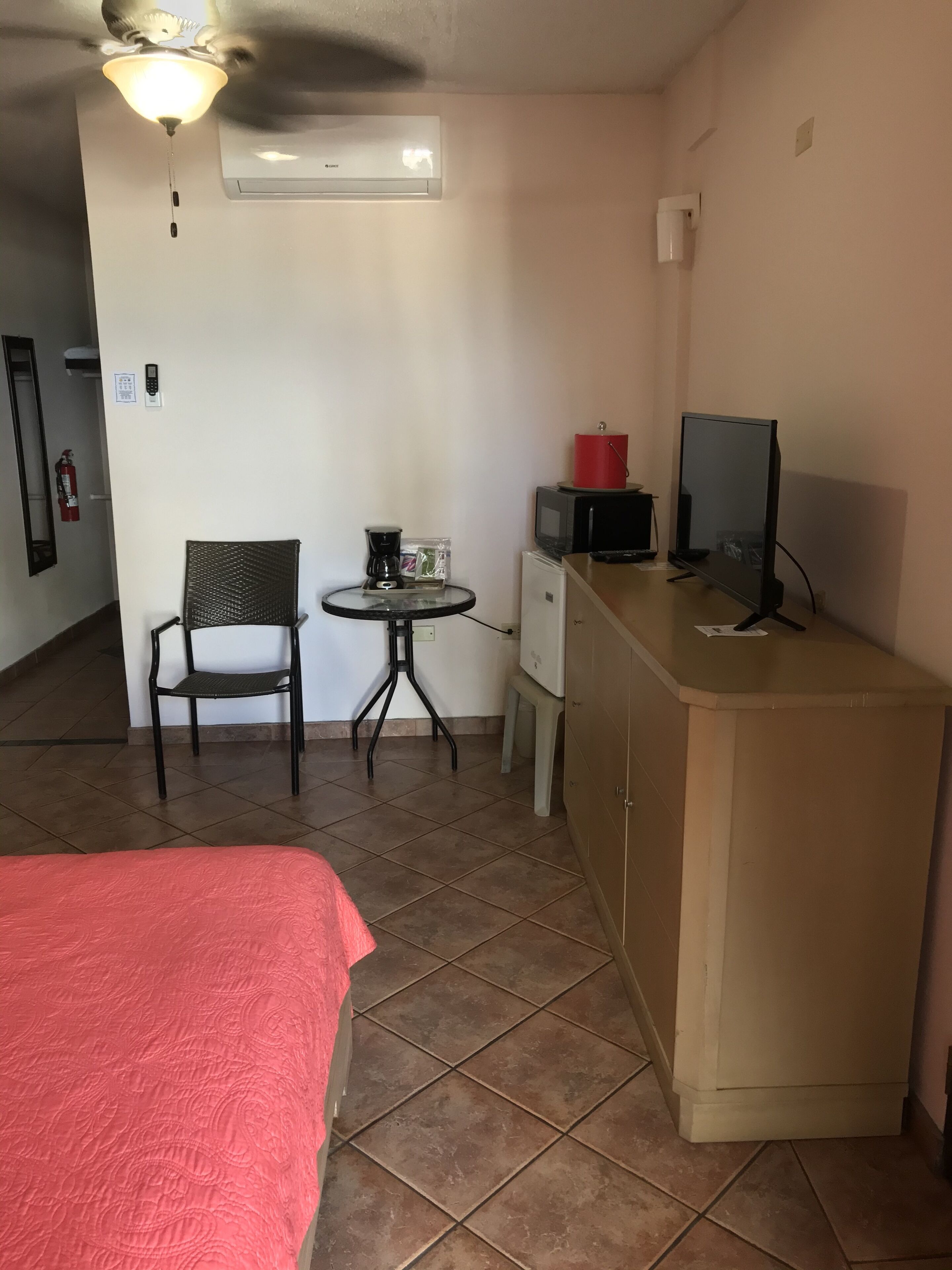 A small, tidy room with a bed, a desk with a computer, a chair, a small table with a coffee maker, and a ceiling fan.