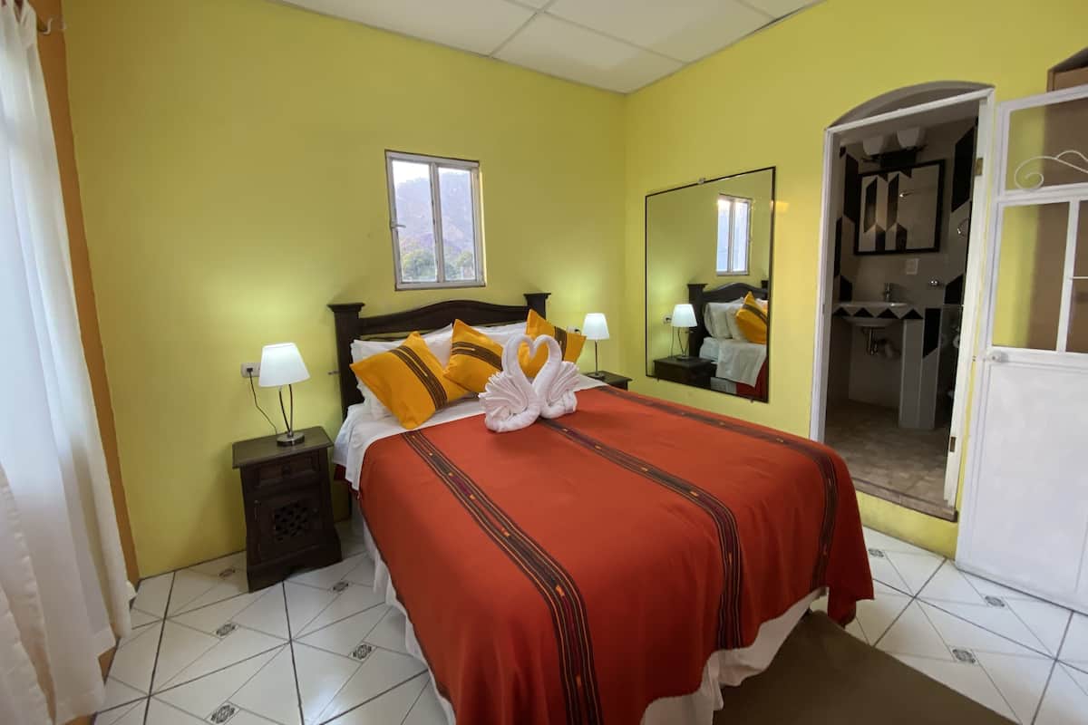 Habitación de hotel con cama, lámparas de noche, ventana y espejo, además de un baño visible a través de una puerta abierta.