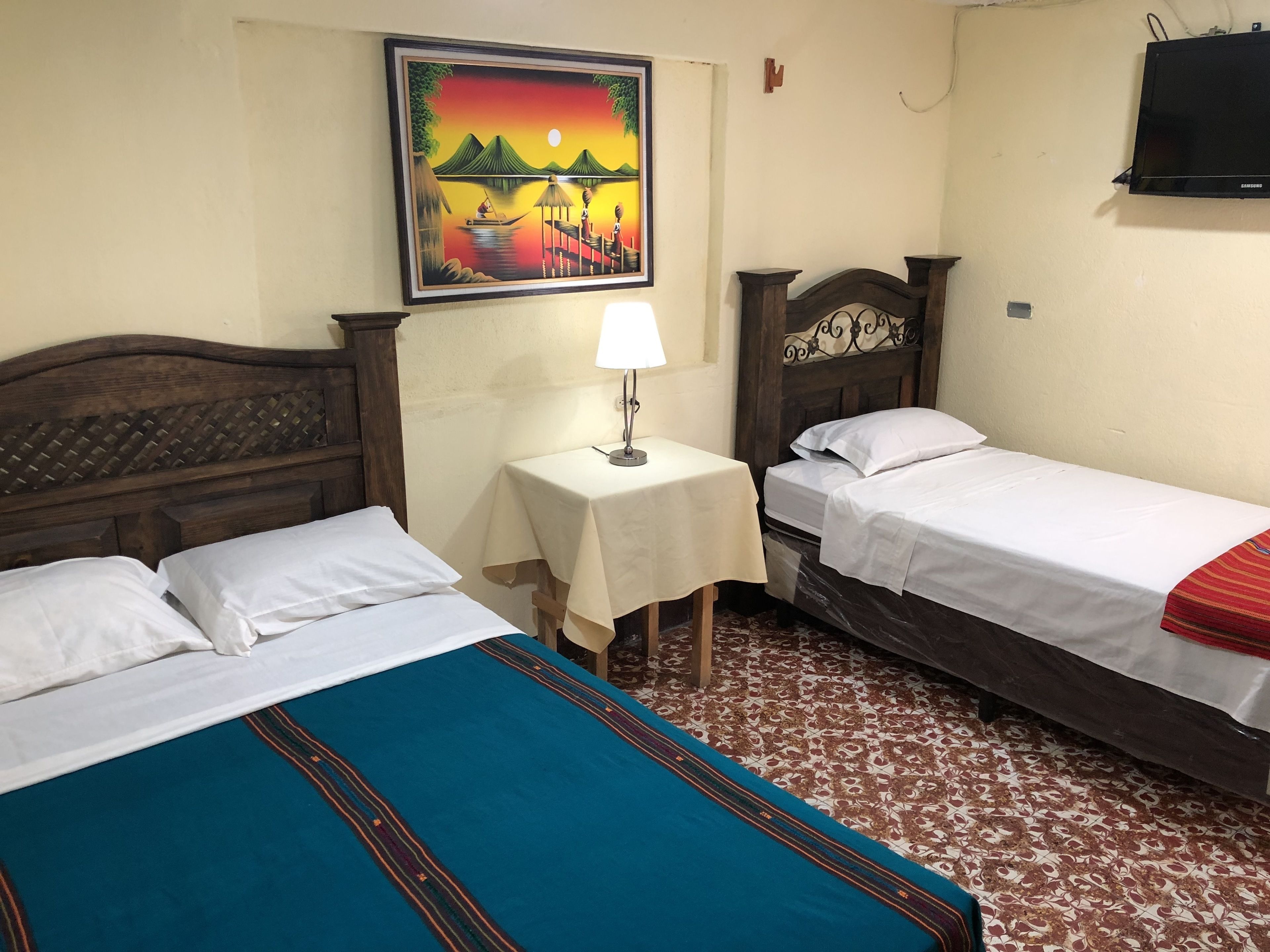 A hotel room with two single beds, a small table with a lamp, a painting on the wall, and a TV mounted on the wall.