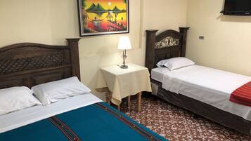 A hotel room with two single beds, a small table with a lamp, a painting on the wall, and a TV mounted on the wall.