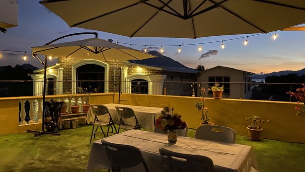 Een terras buiten met een tafel en stoelen, een grote parasol, lichtjes en potplanten.