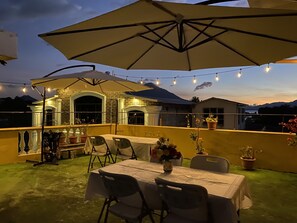Een terras buiten met een tafel en stoelen, een grote parasol, lichtjes en potplanten.