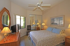 Quarto com cama, escrivaninha com abajur, cadeira, televisão, ventilador de teto e um espelho grande.