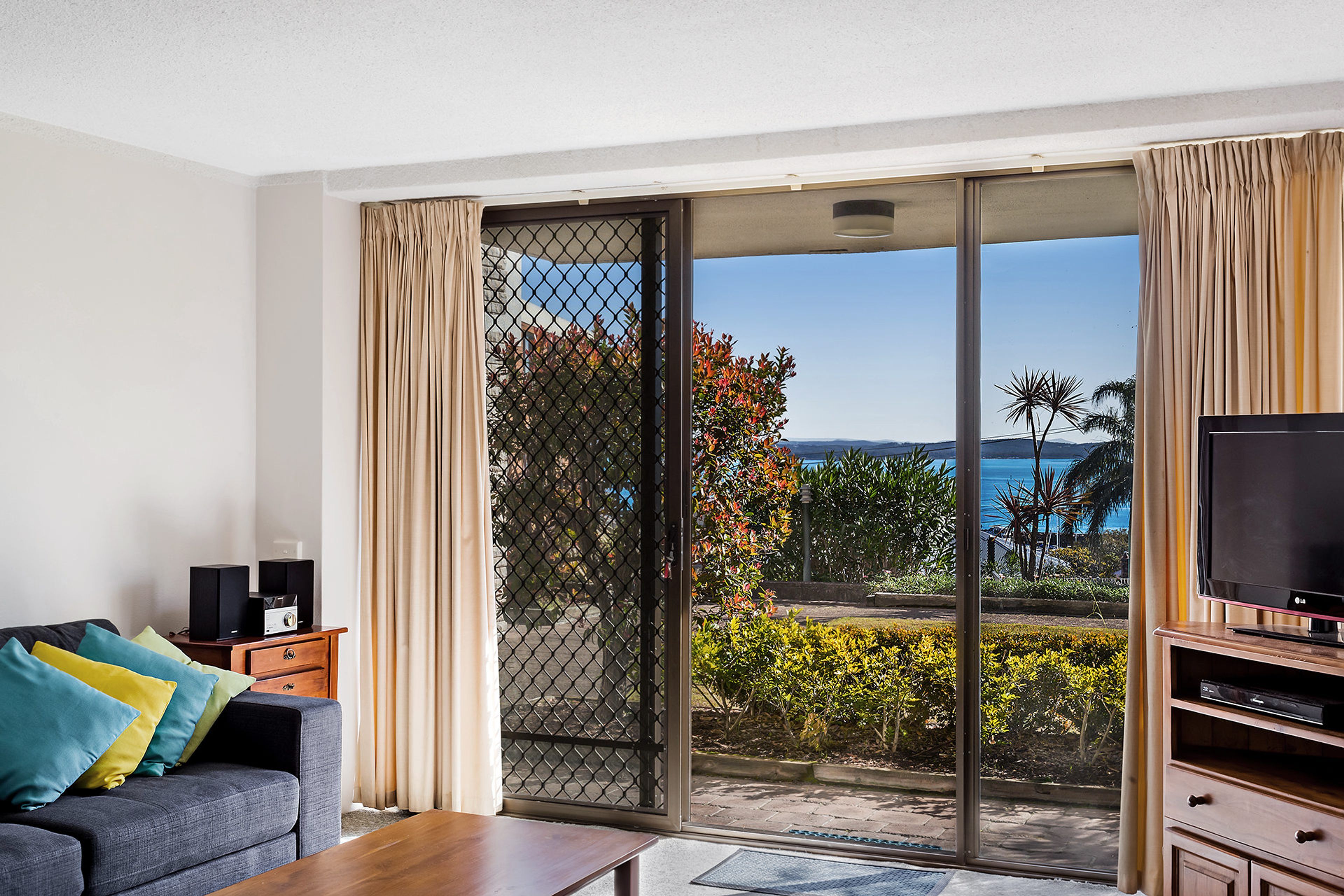 A modern living room with a sofa, television, and speakers, featuring a sliding glass door leading to a balcony with a view of the ocean.