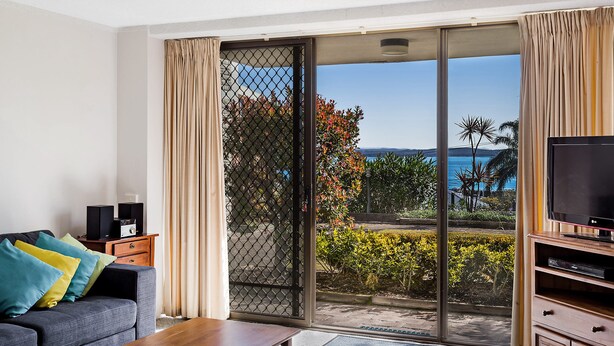 Een moderne woonkamer met een bank, televisie en speakers, met een schuifdeur naar een balkon met uitzicht op de oceaan.