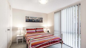 A bedroom with a bed, two bedside tables, a nightstand, a painting on the wall, and a window with blinds. - A ground floor unit, close to town (Nelson Bay)