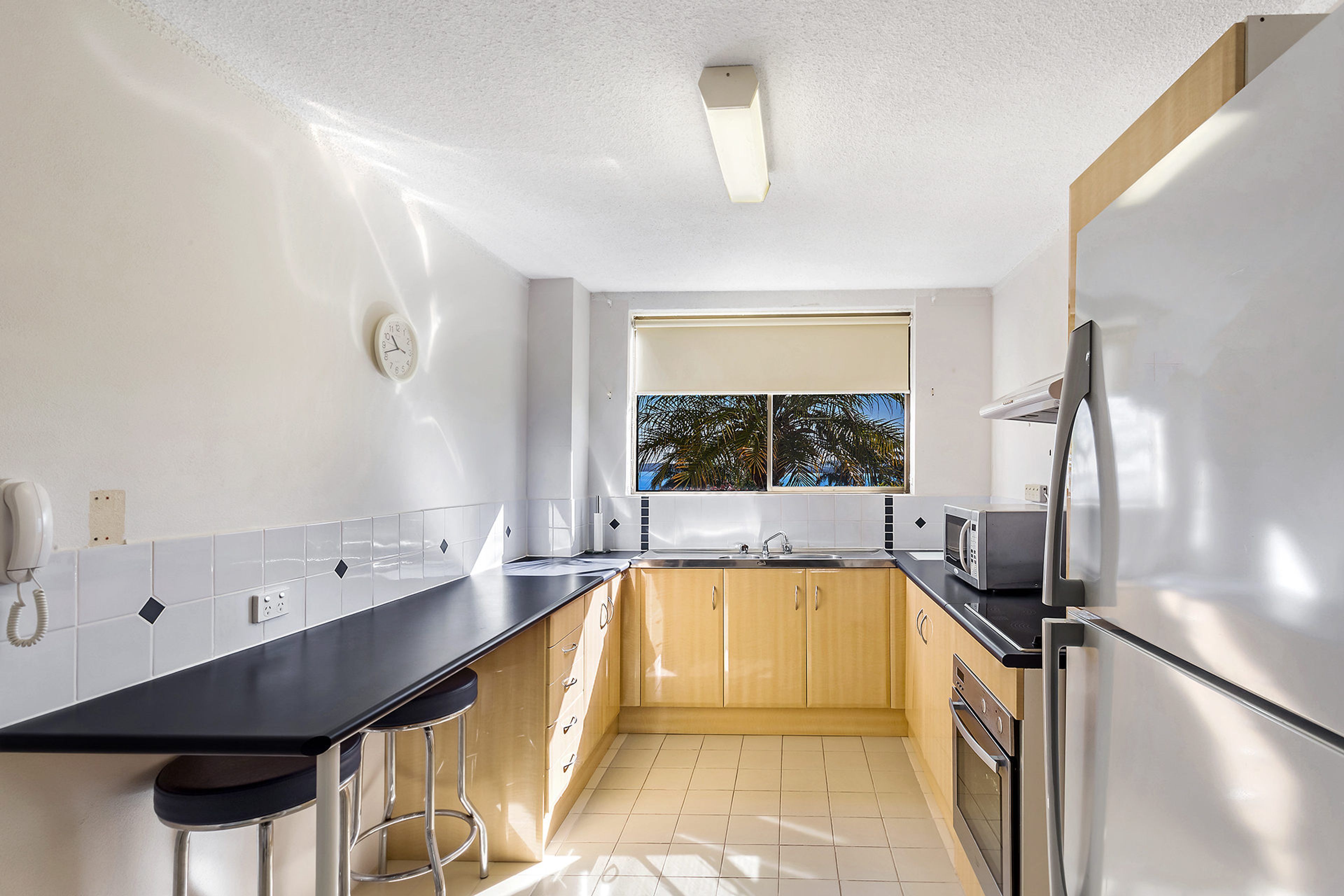 A modern kitchen with a black countertop, wooden cabinets, a stainless steel refrigerator, and a window with a view of palm trees.
