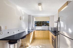 A modern kitchen with a black countertop, wooden cabinets, a stainless steel refrigerator, and a window with a view of palm trees.