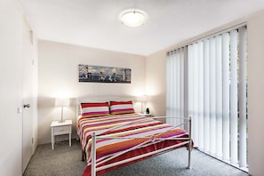 Une chambre à coucher avec un lit, deux tables de chevet, une commode, un tableau au mur et une fenêtre avec des stores.