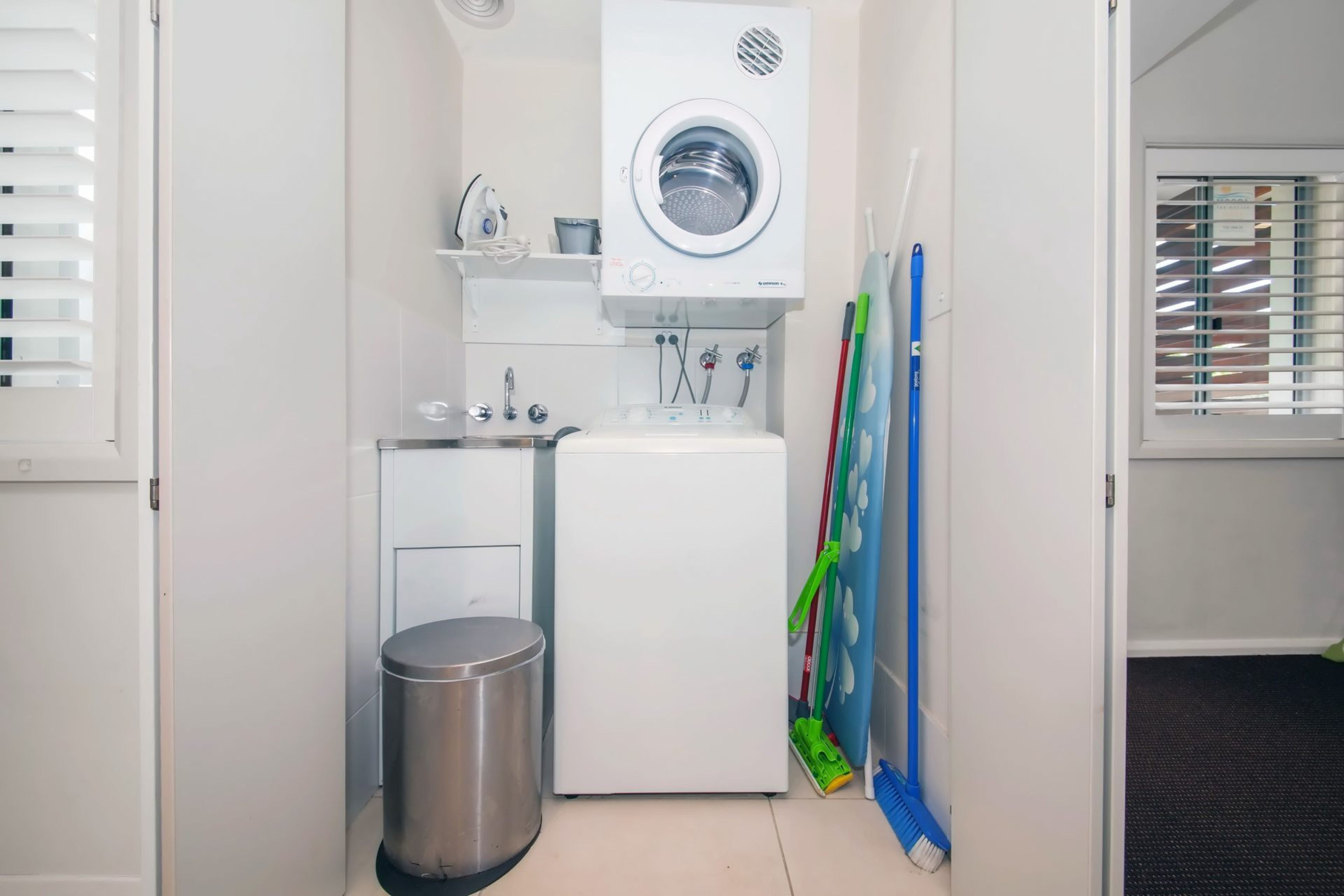 A compact laundry room with a washing machine, a small trash bin, and cleaning tools.