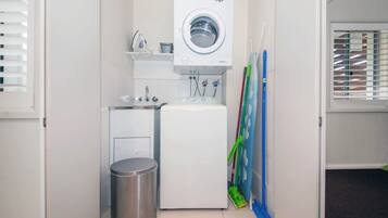 A compact laundry room with a washing machine, a small trash bin, and cleaning tools.