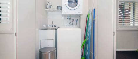 A compact laundry room with a washing machine, a small trash bin, and cleaning tools.