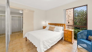 Een slaapkamer met een bed, nachtkastje, houten dressoir, stoel, raam met uitzicht op een bakstenen muur en groen, en een deur naar een badkamer.