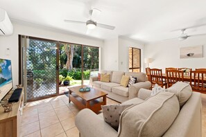A spacious living room with a sliding glass door leading to a garden, a dining area with wooden furniture, and ceiling fans.