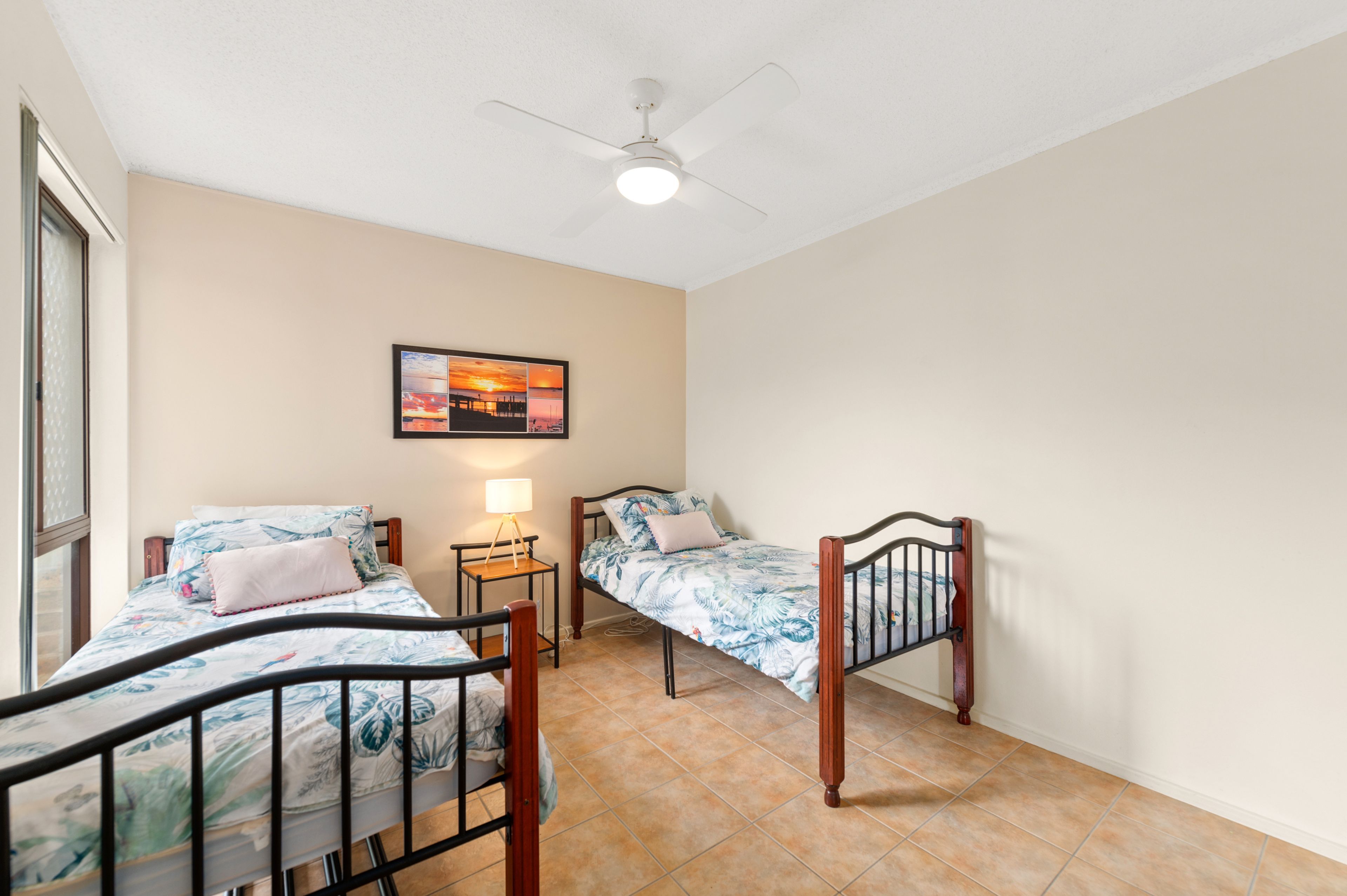 A bedroom with two single beds, a ceiling fan, a nightstand with a lamp, and a framed picture on the wall.