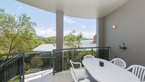 Un balcon avec une table et des chaises blanches, une plante en pot et une vue sur une plage et des collines.