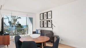 A modern living room with a round wooden table, a grey sofa, and a large window overlooking a balcony with a view of the sea.