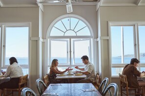 A dining area with a large table and chairs, large windows with a view of the sea, and a ceiling fan.