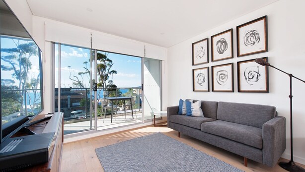A modern living room with a grey sofa, a patterned rug, a piano, and a balcony with a view of the sea.