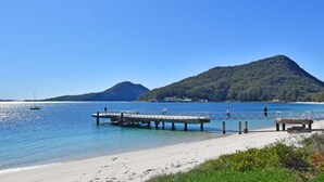 Uma praia com um píer se estendendo sobre águas calmas, céu claro e uma montanha ao fundo.
