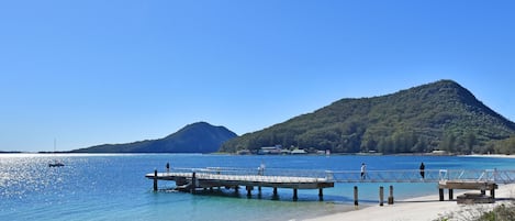 A beach with a pier extending into calm waters, a clear sky, and a mountain in the background.