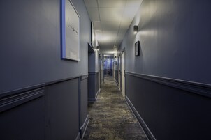 Um corredor comprido e estreito, com piso coberto por carpete, ladeado por paredes com quadros emoldurados e uma parede azul de destaque.