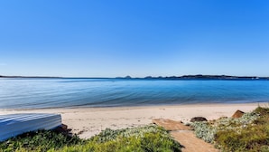 A beach with clear blue water, a sandy shore, and a small path leading to a white structure. - Harbourside, Unit 2/7 Soldiers Point Road - Absolute waterfront unit at Sunrise Beach (Soldiers Point)