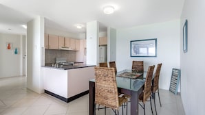A modern kitchen with a central island, a dining area with wicker chairs, and a framed artwork on the wall. - Harbourside, Unit 2/7 Soldiers Point Road - Absolute waterfront unit at Sunrise Beach (Soldiers Point)